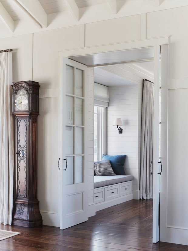 Bright, refined interior showing a wide cased opening with glass-panelled built-in doors leading to a compact window seat nook. The space features white painted wall panelling, shiplap-style lining, built-in bench cabinetry with drawers, flowing curtains, dark timber flooring, exposed white ceiling beams, and an ornate traditional grandfather clock.
