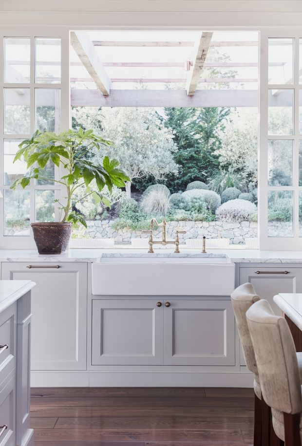 Bright, refined kitchen interior centred on a large white farmhouse apron-front sink beneath a broad garden-facing window. The room features pale grey-white raised-panel cabinetry with dark metal pulls, a light stone benchtop, warm brass-toned tapware, dark timber flooring, and a timber dining or island area with upholstered chairs. Garden greenery and a pergola are visible beyond the glazing.