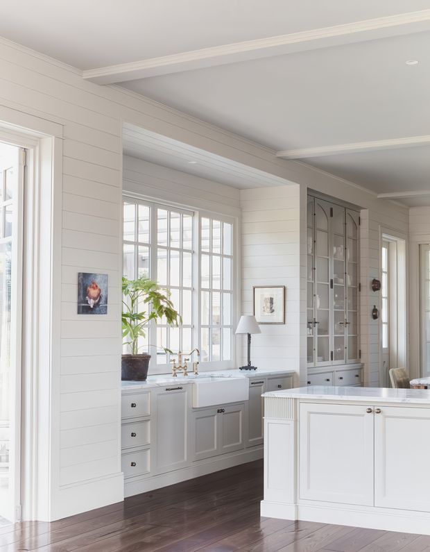 Bright, refined kitchen interior with white horizontal wall panelling, a large multi-pane window, farmhouse-style sink, pale fixed cabinetry, dark timber flooring, and a white island. Glazed built-in display cabinetry and classic trim details add a traditional coastal character.