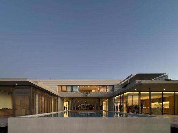 Wide exterior view of a large contemporary flat-roof residence arranged around a central courtyard pool. The low-slung building has extensive floor-to-ceiling glazing, dark framed windows, warm interior lighting, broad roof overhangs, and pale tiled or rendered surfaces. The image is photographed at dusk, with the reflecting pool and courtyard wall in the foreground.