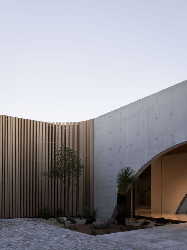 Exterior architectural view of a contemporary courtyard entry, featuring a curved vertical timber-slat screen, a smooth exposed concrete wall with a large arched opening, warm recessed interior lighting, sculptural planting, and pale stone or concrete paving.