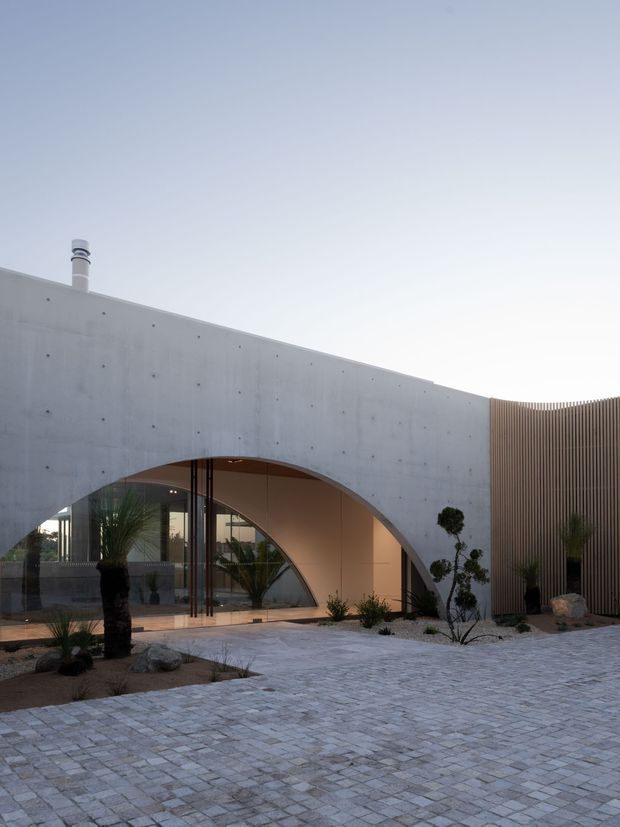 Exterior view of a minimalist contemporary building with a pale concrete facade, dramatic curved arch opening, extensive glazing, and a tall vertical timber-screen wall. A paved courtyard with sparse sculptural landscaping, rocks, palms, and clipped trees forms the foreground.