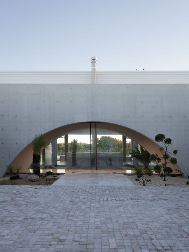 Minimalist modern building exterior with a broad concrete facade, a prominent arched recessed entry, full-height glass doors, and a pale stone-paved forecourt. Sparse sculptural landscaping with palms and carefully pruned trees frames the entrance, while a flat roof and rooftop flue reinforce the geometric architectural form.