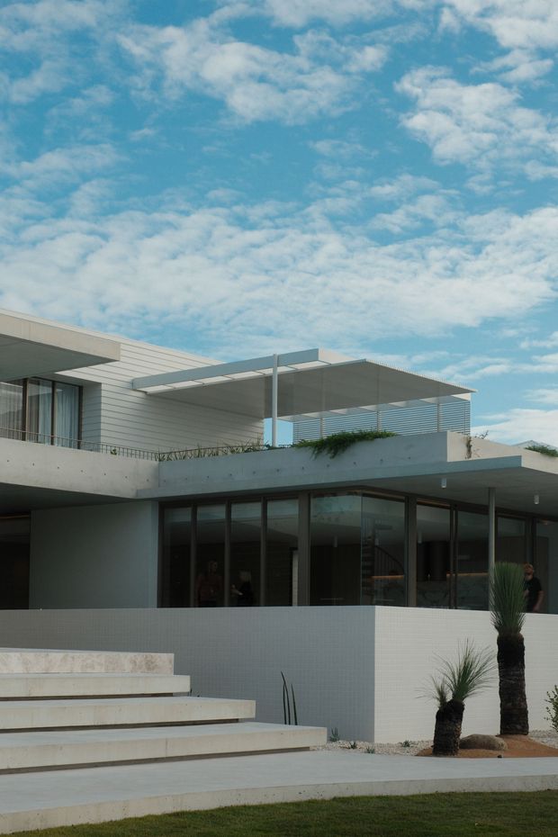 Modern multi-level residential house with a flat roof, projecting concrete terraces, pale horizontal cladding, extensive dark-framed glazing, and a rooftop terrace with a lightweight pergola and horizontal balustrade. Broad concrete steps lead up to the house, with sparse architectural landscaping and sculptural palm-like plants in the foreground. The image is photographed from outside under a bright partly cloudy sky.