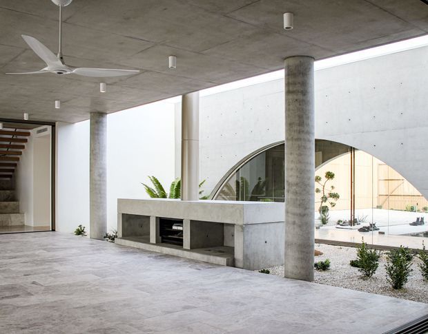 A minimalist concrete courtyard or covered outdoor living area with a broad tiled floor, exposed concrete ceiling, cylindrical concrete columns, and a built-in concrete bench or barbecue counter. A large arched opening frames a bright gravel courtyard and planting beyond. A staircase and white ceiling fan are visible at the left.