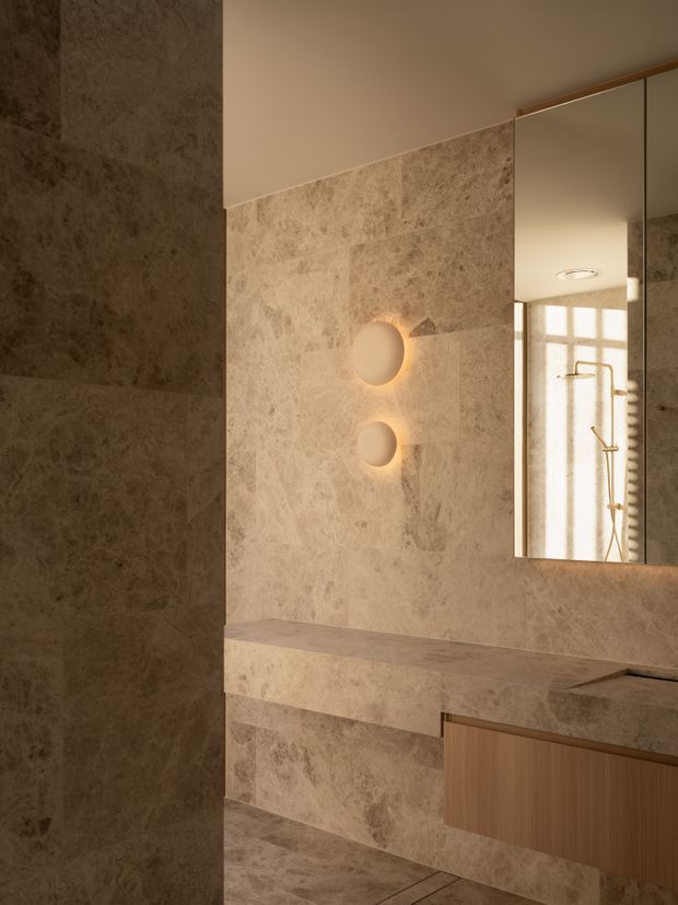 Warm, refined bathroom interior featuring full-height beige natural-stone wall and floor surfaces, a matching stone vanity benchtop and integrated shelf, timber vanity cabinetry, round illuminated wall lights, and a large mirrored cabinet reflecting a glass shower with brass-toned fixtures.