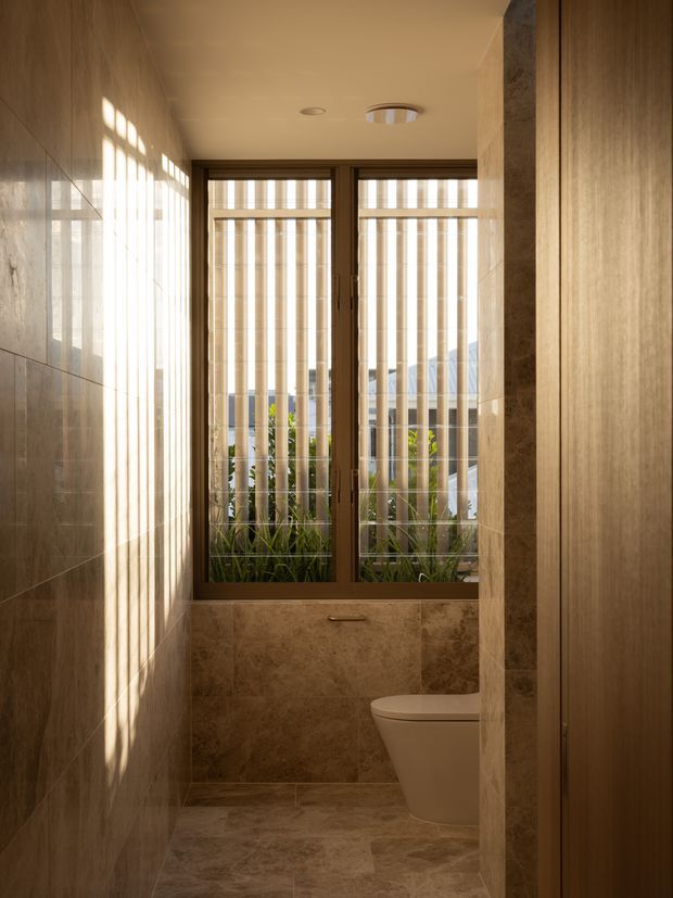 A narrow, warm-toned bathroom with full-height beige-brown stone-look wall and floor tiles, a concealed or partially visible toilet, and a large timber-framed window fitted with vertical blinds. Strong sunlight creates striped shadows across the tiled walls and floor. Fixed timber cabinetry or a door is visible on the right, and planting can be seen beyond the window.