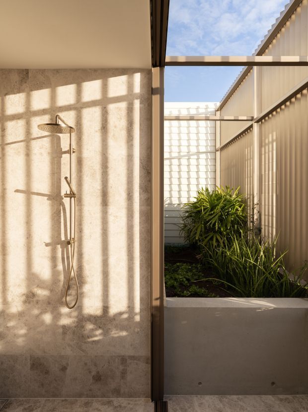 A contemporary open-air bathroom shower is shown beside a narrow planted courtyard. The shower has a large natural stone-tiled wall and floor, brushed metal fixtures, and a full-height glazed opening framed in dark metal. Warm sunlight casts strong geometric shadows across the stone and adjoining vertical-panel enclosure, with lush planting in a raised concrete planter outside.