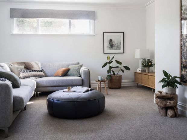 A bright, softly styled living room with a large light-grey sectional sofa, black round ottoman, neutral carpet, white walls, and a timber mid-century-inspired sideboard. Potted plants, framed artwork, cushions, and a horizontal window with a grey roller blind add warmth and texture. The room has a calm, contemporary feel with natural light and restrained neutral tones.