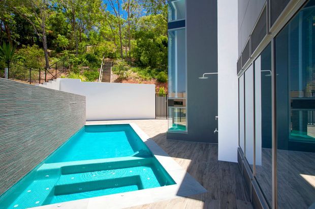 A contemporary residential courtyard viewed beside a modern building, featuring a narrow stepped turquoise swimming pool with integrated spa-like seating, pale stone paving, a textured rendered privacy wall, dark vertical fencing, and extensive floor-to-ceiling glazing with strong reflections. A landscaped hillside and stairway rise behind the courtyard.