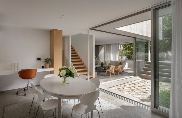 Bright, refined open-plan dining and living interior with a round white dining table and contemporary chairs in the foreground. Full-height sliding glazing opens toward a landscaped courtyard and an adjoining lounge area, while a sculptural timber staircase rises beside a pale vertical feature wall. The room has a restrained neutral palette, abundant natural light, and clean modern detailing.