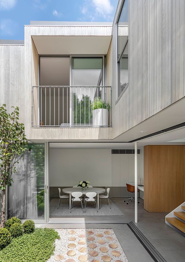 A bright contemporary multi-level courtyard interior/exterior view. The image looks across a landscaped courtyard toward an open-plan dining area, with a sheltered upper balcony, extensive glazing, pale vertical cladding, slim metal balustrades, a timber pivot door, and a stair ascending at right. The dining space contains a round table and chairs, while rounded planting and stepping stones define the foreground.
