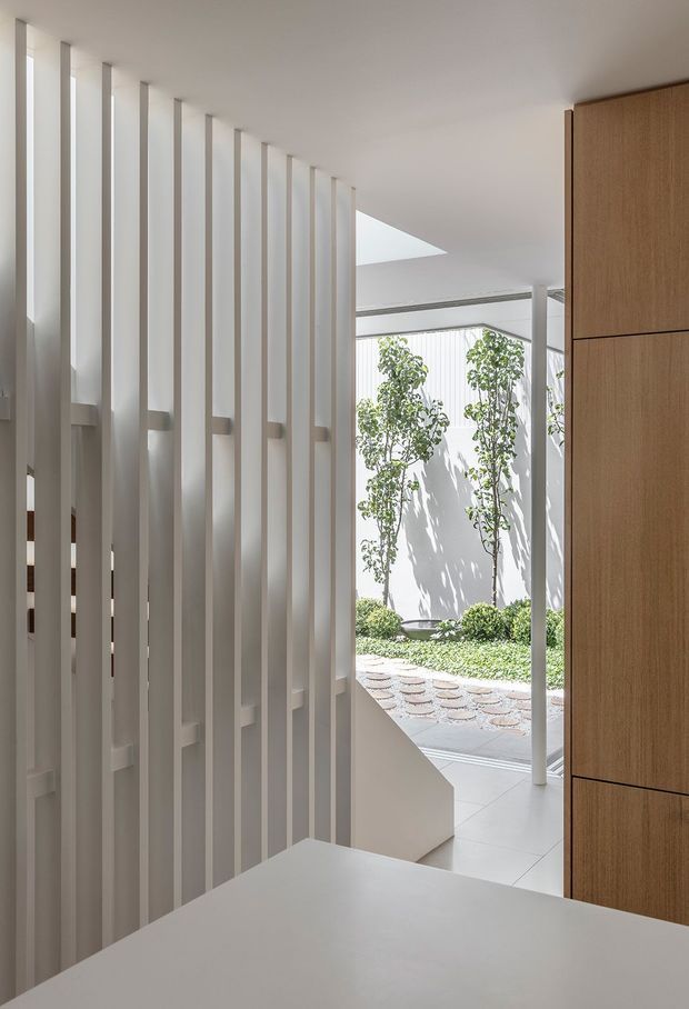 Bright minimalist interior transition space with a white sculptural vertical-slat screen, pale tiled flooring, and a large light-oak built-in cabinet. The view opens toward a planted courtyard through extensive glazing, with a skylight or ceiling opening bringing in daylight. A white foreground surface partially obscures the lower frame.