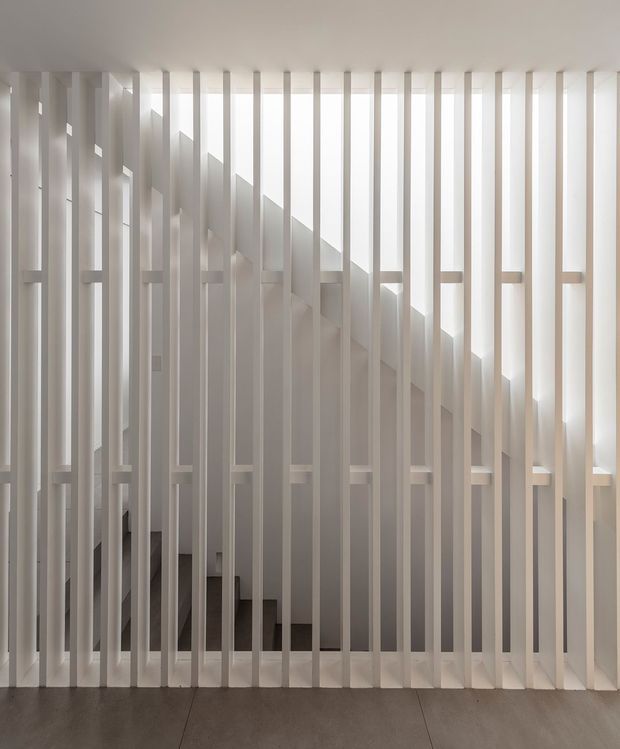 A minimalist interior view of a stairway screened behind closely spaced vertical white slats. The slatted partition extends from floor to ceiling, allowing filtered daylight to cast strong linear shadows across the stair enclosure and walls. A dark stair flight and pale flooring are visible behind the screen.
