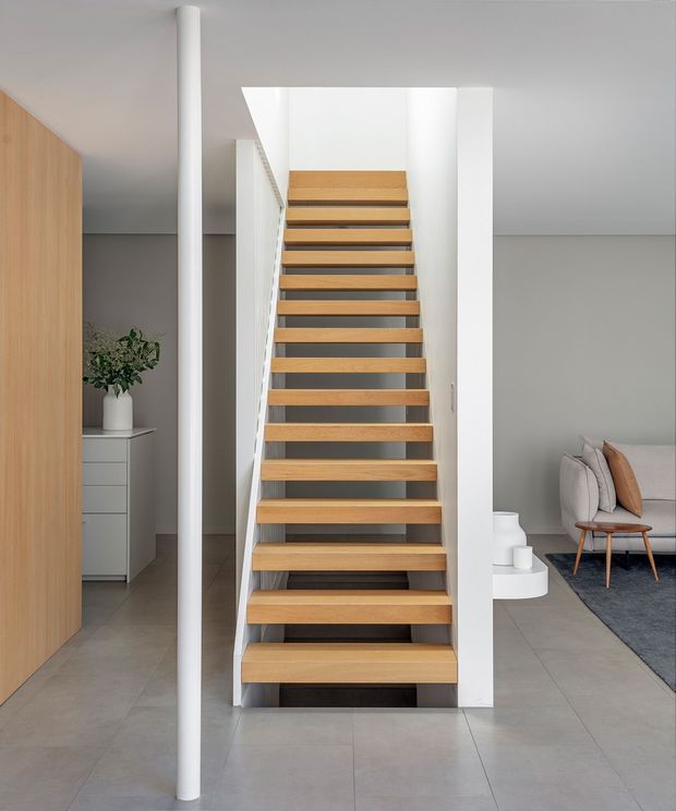 A bright, minimalist interior focused on a straight floating staircase with warm timber treads, white side walls, and a slim white structural column. The stairway connects an open-plan living area, with light grey tiled flooring, pale grey walls, timber cabinetry, and a partially visible lounge with a grey sofa and small timber-framed chair. Clean lines, concealed detailing, and abundant neutral tones create a refined contemporary setting.