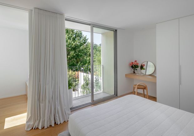 Bright minimalist bedroom with a low-profile white bed, pale timber flooring, full-height sliding glass doors opening to a balcony, sheer floor-to-ceiling curtains, and streamlined white built-in wardrobes. A simple timber console with a round mirror and small floral arrangement sits against the wall. The room has a clean, airy, understated contemporary appearance.