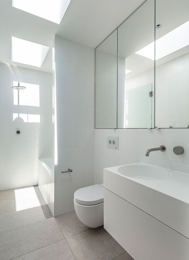 Bright minimalist bathroom with a walk-in shower, wall-hung toilet, freestanding-style bathtub, and floating vanity. The space is predominantly white with large mirrored cabinetry, pale tiled walls, a light grey tiled floor, frameless glass, and multiple skylights providing abundant natural light.