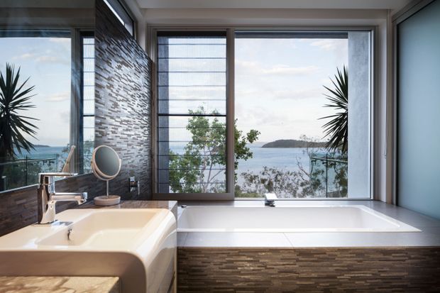 A bright, modern bathroom with a freestanding-style built-in bathtub positioned beside expansive corner glazing overlooking a coastal bay. The room features a double vanity with chrome tapware, a large mirror, textured stacked-stone wall and bath-front cladding, and dark-framed windows. Tropical plants and the water view add a relaxed coastal atmosphere.