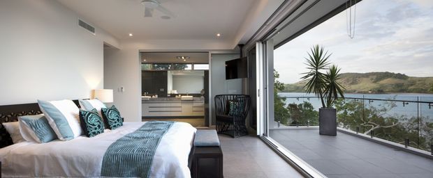 Bright contemporary bedroom with a bed in the foreground, opening directly onto a tiled balcony through large full-height sliding glass doors. A compact kitchen or living area is visible beyond the bedroom, while the balcony overlooks a leafy hillside and water. The room uses a restrained neutral palette with blue-green textiles, streamlined built-in cabinetry, recessed lighting, and a large potted plant outside.