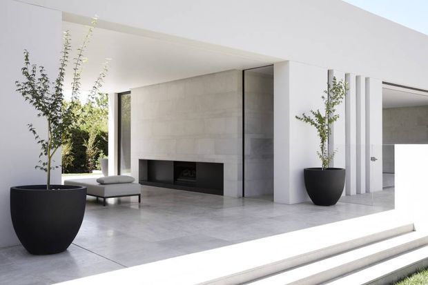 Bright minimalist contemporary outdoor-covered living area with a large open fireplace, pale stone or concrete feature wall, expansive white structural forms, and broad pale tiled or concrete flooring. Large black planters with slender trees flank the space, while a low lounge chair sits near the opening. Steps lead up to the covered terrace, with a garage-like opening visible at right.
