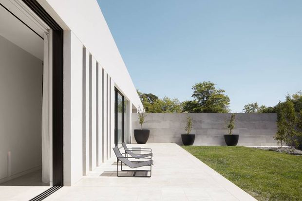 A minimalist modern courtyard terrace beside a stark white, single-storey building. The facade features repetitive vertical fins and large glazed openings, with two reclining chairs placed on a pale paved terrace. A concrete boundary wall, dark planters, lawn, and mature trees complete the quiet outdoor setting.