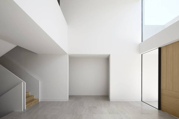 Minimalist interior view of a bright, open stair hall or landing. White geometric walls and soffits frame a timber staircase and a recessed wall opening, while a tall black-framed glazed door or partition and a warm timber-panelled doorway appear on the right. The pale grey floor and abundant daylight create a clean, contemporary atmosphere.