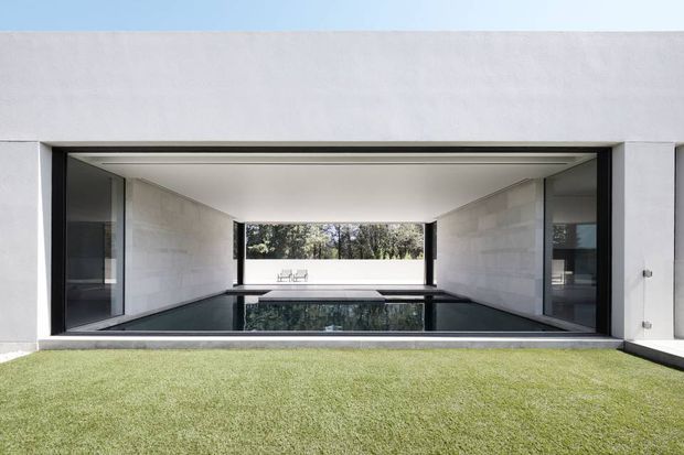 A minimalist modern architectural view from a lawn into a long, open-sided courtyard or covered outdoor room. White rendered walls and pale stone-clad side walls frame a dark reflective rectangular pool or water feature, with large black-framed glazed openings and a narrow rear view toward trees. The composition is geometric, symmetrical, and strongly connected to the outdoors.