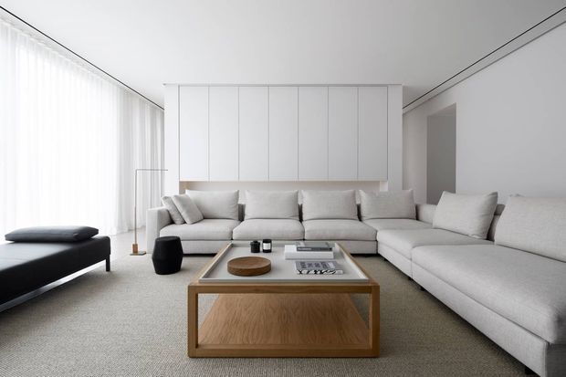 A bright, minimalist living room with a large light-grey sectional sofa, matching ottoman, and a low rectangular timber-and-glass coffee table. Floor-to-ceiling sheer curtains line the left wall, while seamless white built-in cabinetry or wall panelling forms a clean feature behind the sofa. The room has a restrained neutral palette, concealed detailing, and a calm contemporary appearance.
