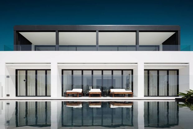 Front-on view of a refined modern multi-level residence with a crisp white facade, dark upper-storey framing, extensive floor-to-ceiling glazing, and a covered outdoor terrace furnished with loungers. A reflective swimming pool occupies the foreground, mirroring the architecture. The composition is symmetrical and minimalist, with a deep blue sky and sparse planting at the edge.