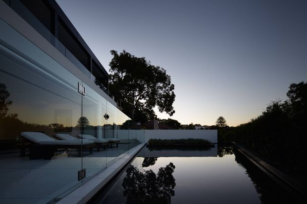 Twilight exterior view of a sleek contemporary residence with extensive floor-to-ceiling glazing, a glass balustrade, and a long reflective rectangular swimming pool. Minimal outdoor loungers are visible behind the glazing, while trees and the darkening sky frame the property.