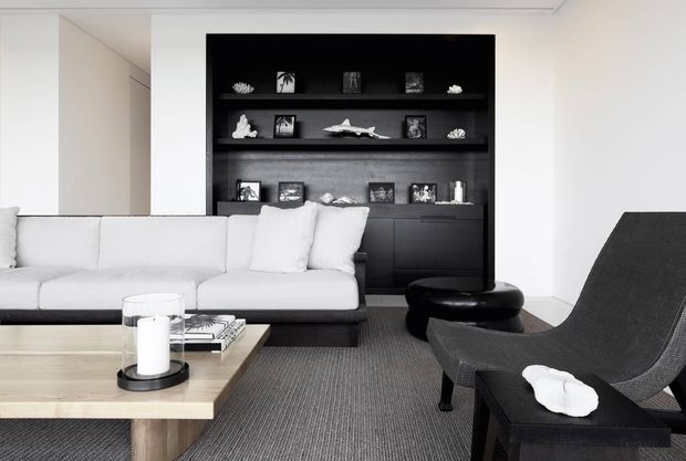 A refined monochrome living room with a large white sectional sofa, low pale timber coffee table, dark built-in shelving and cabinetry displaying decorative objects, and a sculptural black lounge chair with matching ottoman. The space has a clean, restrained contemporary composition with strong black-and-white contrast, soft natural light, and a textured grey carpet.