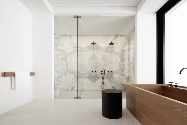 Bright contemporary bathroom with a large frameless glass walk-in shower, dramatic white marble-look wall tiling, pale floor tiles, and a freestanding timber bathtub beside a black-framed window. Minimal black fixtures and a black cylindrical stool add contrast.