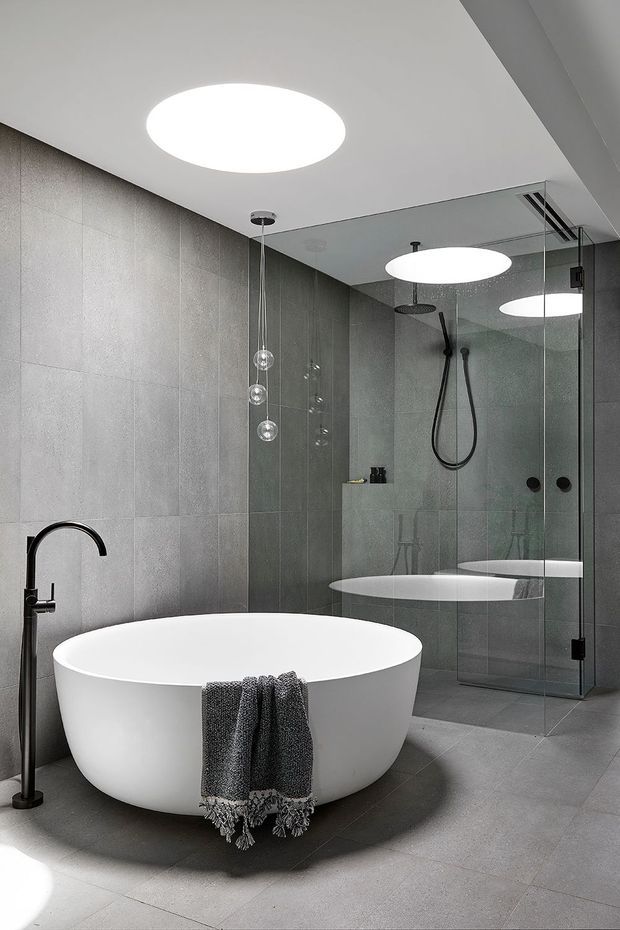 A contemporary bathroom featuring a freestanding white oval bathtub, wall-mounted black floor-standing faucet, large grey tiled walls and floor, and a frameless glass walk-in shower with dark fittings. Round recessed ceiling lights and pendant globe lights create a clean, refined atmosphere.