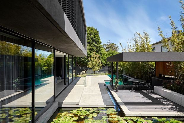 Contemporary multi-level residential courtyard viewed from an outdoor terrace, with extensive floor-to-ceiling glazing, a concrete upper volume, linear reflecting ponds with water lilies, pale paved walkways, and a covered outdoor dining and lounge area. Dense landscaping provides privacy around the pool and courtyard.