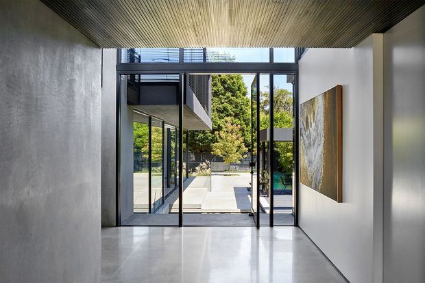 A refined modern entry hall with polished light-grey flooring, smooth white and concrete-textured walls, and a dark linear slatted ceiling. Full-height black-framed glazing and an open glass door connect the hall to a bright paved courtyard and pool area beyond. A large abstract artwork adds a warm natural accent to the otherwise minimalist interior.