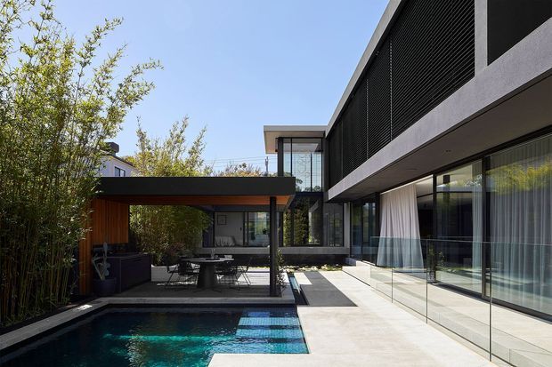 Contemporary two-storey residential exterior viewed from a landscaped courtyard, with a dark horizontal facade, extensive floor-to-ceiling glazing, a covered outdoor dining area, and a compact swimming pool with paved terrace. Timber screening and dense bamboo planting soften the strong geometric architecture.