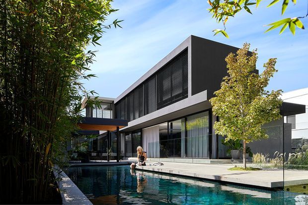 Exterior view of a large contemporary two-storey residence centred around a long swimming pool. The building has crisp rectilinear volumes, dark rendered walls, extensive floor-to-ceiling glazing, horizontal louvres, a projecting upper level, and a pale paved pool terrace. Mature bamboo and landscaped planting frame the foreground, while a transparent pool fence runs along the water.