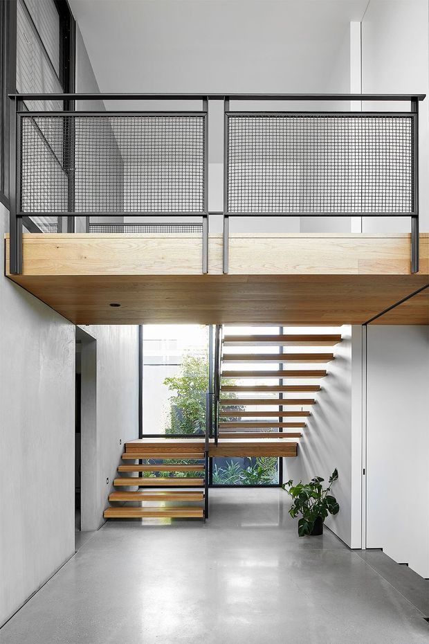 A bright, double-height interior centered on a sculptural open-riser staircase. Floating timber stair treads and a matching timber mezzanine bridge are supported by dark metal framing, with mesh-panel balustrades above. Smooth pale grey concrete flooring and white walls create a clean, minimalist setting, while large windows bring in daylight and views of planted greenery.