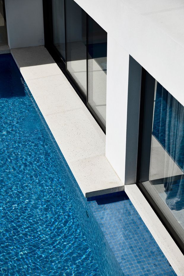A close architectural view of a narrow swimming pool running alongside a modern white building. The pool edge has a pale speckled concrete or stone coping, with blue mosaic tiles visible on a submerged step or ledge. Full-height dark-framed glazing reflects the water and reveals curtains inside, while sharp white geometric wall planes create a clean, high-contrast composition.