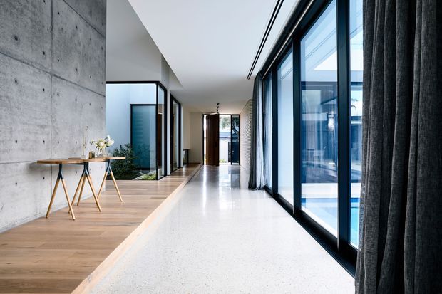 A refined contemporary interior corridor with a long open circulation space, polished white terrazzo-style flooring, warm timber flooring to one side, exposed raw concrete walls, and extensive black-framed floor-to-ceiling glazing. Dark full-height curtains line the glazing, with a small timber console table and decorative objects against the concrete wall. A glimpse of a blue swimming pool is visible outside through the windows.