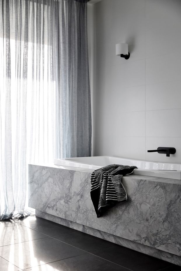 A refined contemporary bathroom featuring a freestanding or built-in white bathtub with a grey marble-clad bath surround, large pale wall tiles, dark floor tiles, and matte black tapware and wall light. Full-height sheer grey curtains filter strong natural light from a tall window, creating a calm monochromatic atmosphere.