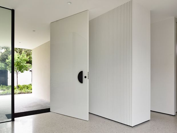 Bright minimalist interior entry with a large white pivot door, black door hardware, floor-to-ceiling black-framed glazing, and a prominent white vertically fluted feature wall. A terrazzo-like light floor and views to a landscaped exterior reinforce the clean contemporary design.