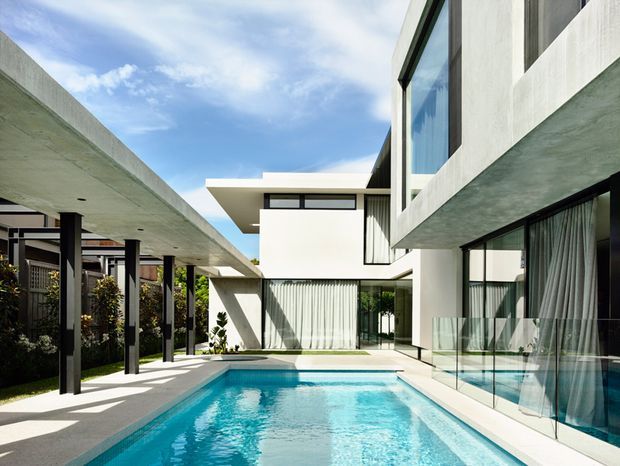 A bright contemporary multi-level residence organized around a rectangular swimming pool and courtyard. White rendered volumes, extensive floor-to-ceiling glazing, projecting flat roof slabs, dark slender structural posts, and a covered outdoor walkway create a clean geometric composition. The pool is bordered by pale paving and a frameless glass balustrade, with landscaped planting along the left side.