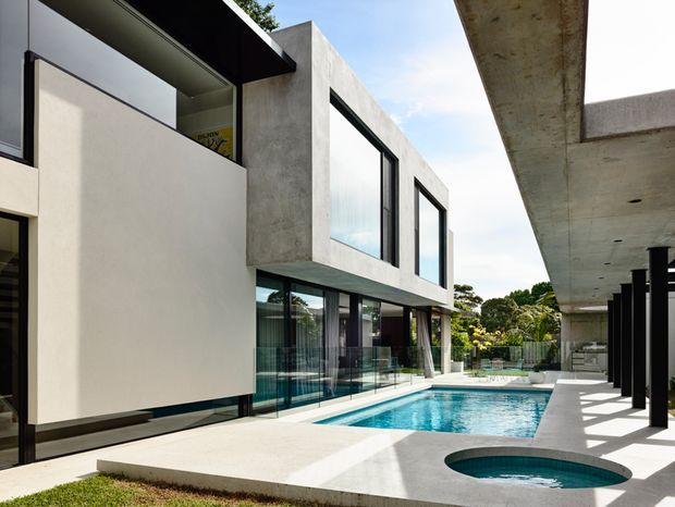 Contemporary multi-level residence arranged around a long swimming pool and circular spa. The exterior features strong rectilinear concrete forms, extensive floor-to-ceiling glazing with dark frames, pale rendered walls, and broad cantilevered roof slabs. A covered outdoor terrace with dark structural posts runs alongside the pool, creating a clean, resort-like courtyard setting.