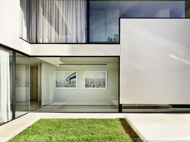 A contemporary multi-level interior courtyard framed by extensive floor-to-ceiling glazing. The image shows a minimalist white and grey architectural composition, with a grass courtyard in the foreground, a glazed living or circulation space beyond, and an upper-level balcony or room with sheer curtains. Clean horizontal lines, recessed lighting, large glass panels, and built-in architectural forms dominate the scene.