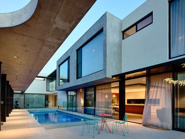 Contemporary multi-level residence arranged around a long narrow swimming pool and paved courtyard. The building features strong rectilinear forms, exposed concrete soffits and walls, extensive dark-framed glazing, projecting upper-level volumes, and an open-plan illuminated interior with curtains and built-in joinery visible through the glass. Bright blue sky and minimal landscaping frame the architectural composition.