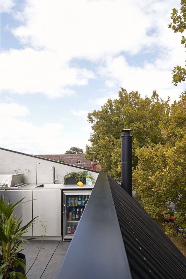 A compact rooftop outdoor kitchen or entertaining area beside a dark metal-clad roof edge. The space includes stainless-steel cabinetry, a sink with tap, tiled flooring, a built-in drinks refrigerator, and a tall black flue or chimney. Trees and neighbouring rooftops are visible beyond the parapet under a bright partly cloudy sky.