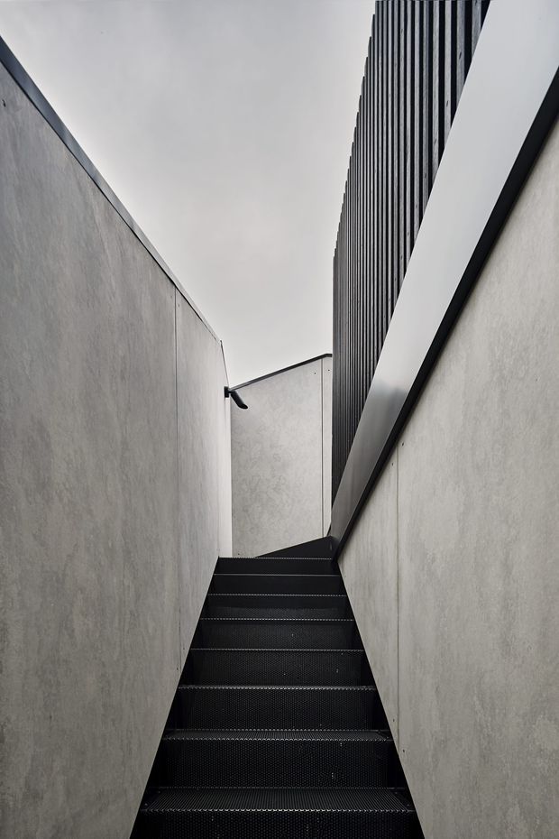 A narrow exterior stairway rises between tall pale grey rendered concrete or plaster walls. The stairs and upper vertical screening are black metal, with a strongly linear, minimalist composition and an overcast sky visible above.