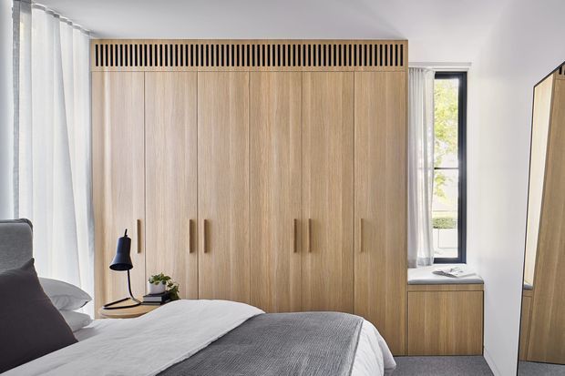 A bright, contemporary bedroom featuring a large built-in timber wardrobe with vertical slatted ventilation detailing at the top, integrated handles, and matching low cabinetry beneath a narrow window. The bed is dressed in white and grey bedding, with sheer curtains, a simple bedside lamp, and a full-height mirror at the right.