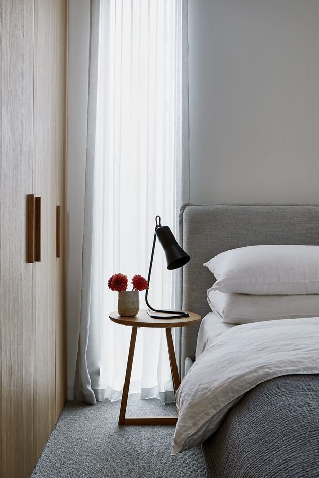 A softly lit bedroom corner with a grey upholstered bed, layered white bedding, a slim wooden bedside table, black task lamp, and a small vase of red flowers. Full-height sheer curtains provide diffused natural light, while light timber wardrobe cabinetry lines the left side. The room has a calm, refined contemporary appearance with neutral textures and warm timber accents.